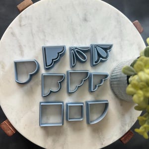 May include: A collection of light blue corner-shaped cookie cutters in various decorative designs. The cutters are arranged on a white marble surface, with a few wooden accents and a small metal container with greenery.