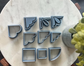 May include: A collection of light blue corner-shaped cookie cutters in various decorative designs. The cutters are arranged on a white marble surface, with a few wooden accents and a small metal container with greenery.