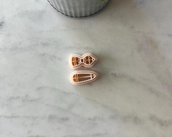 May include: Two beige plastic cookie cutters. One is shaped like a bow tie, and the other is shaped like a pencil. The cookie cutters are on a white marble surface. A metal pot with green plants is in the background.