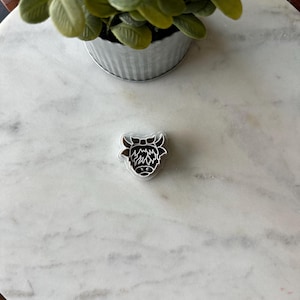 May include: A silver metal cookie cutter shaped like a Highland cow with a black outline and a white face.