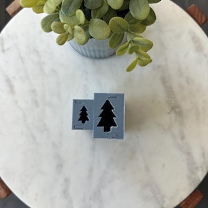 May include: Two gray plastic cookie cutters shaped like Christmas trees. The larger cutter is in the background and the smaller cutter is in the foreground.