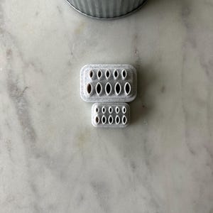 May include: Two gray, rectangular cookie cutters with oval cutouts. The larger cutter has two rows of six cutouts, and the smaller cutter has two rows of five cutouts. The cutters are on a marble surface.
