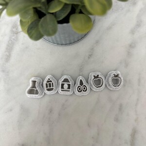May include: A collection of six gray plastic cookie cutters in various shapes, including a beaker, house, gift, scissors, and two apple shapes. The cutters are arranged in a row on a white marble surface, with a small potted plant in the background.