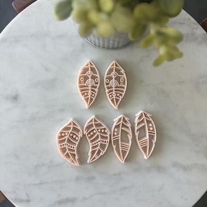 May include: A collection of six leaf-shaped cookie cutters. The cutters are light brown with white interior details, featuring various geometric and feather-like designs. They are arranged on a white marble surface.