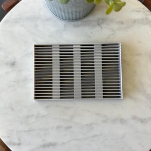 May include: A rectangular, silver-colored metal grate with evenly spaced horizontal bars. The grate is set on a white and gray marble surface, with a small potted plant in the background.