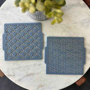 May include: Two blue-grey decorative stencils with intricate cut-out patterns. One features a fan-like design, the other a floral pattern. The stencils are square with small tabs on the sides. The stencils are on a white marble surface.