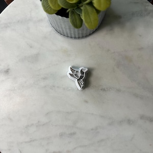 May include: A silver cookie cutter in the shape of a bird on a white marble surface.