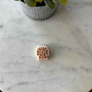 May include: A light brown, flower-shaped cookie cutter with a detailed design. The cutter is made of plastic and sits on a white marble surface. A small, silver metal pot with a green plant is in the background.