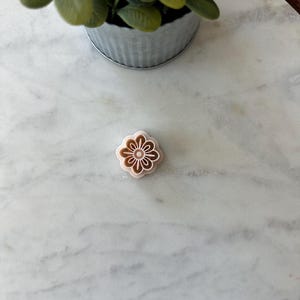 May include: A small, flower-shaped cookie cutter with a light brown exterior and a darker brown interior design. The cutter is centered on a white marble surface, with a small potted plant in the background.