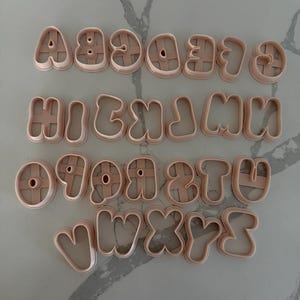 May include: A complete set of twenty-six light pink plastic alphabet cookie cutters arranged in four rows. Each letter features a rounded, bubble-style font with a thick outer frame and a central crossbar or support structure for structural integrity.