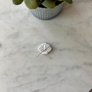 May include: A white seashell-shaped cookie cutter with a thin, straight metal pin piercing through it. The cutter is on a white marble surface, with a small potted plant in the background.
