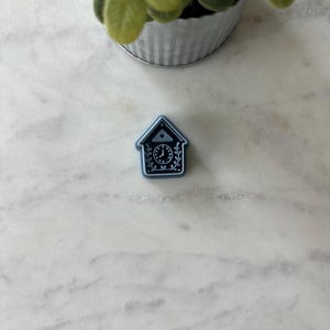 May include: A small, blue and white cuckoo clock-shaped pin. The pin features a clock face with decorative leaves and a house-shaped outline. The pin is set against a white marble surface.