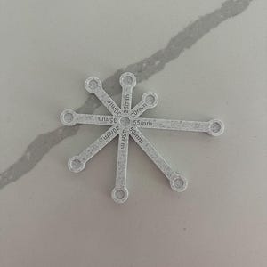 May include: A light gray, star-shaped tool with multiple arms, each labeled with millimeter measurements: 20mm, 25mm, 30mm, 35mm, 40mm, and 55mm. The tool has small circular holes at the end of each arm.