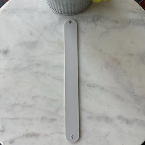 May include: A light gray nail file with a textured surface and a small hole at the top. The number 1 is printed at the bottom of the file. The file is shown on a white marble surface.