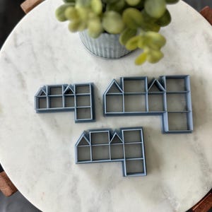 May include: Three light blue house-shaped cookie cutters in various sizes are arranged on a white marble surface. The cutters have rectangular and triangular sections, resembling miniature buildings. A small metal pot with green foliage is in the background.