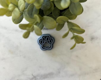 May include: A small, dark blue paw print-shaped object with a black outline. The paw print is centered on a white marble-patterned surface, with green leafy plants in the background.
