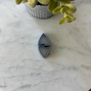 May include: A light blue cookie cutter in the shape of a stylized leaf or shield. The cutter is made of plastic and is lying on a white marble surface. A small metal pot with green plants is in the background.