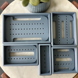 May include: Five gray plastic drawer organizers with different compartments. The organizers are on a white marble surface.