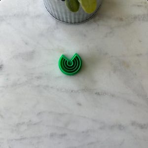 May include: Green plastic cookie cutter shaped like a crescent moon with concentric circles.