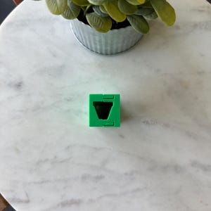 May include: A bright green, geometric cube with a V-shaped cutout. The cube is made of plastic and has a simple, modern design. The cube is sitting on a white marble surface.