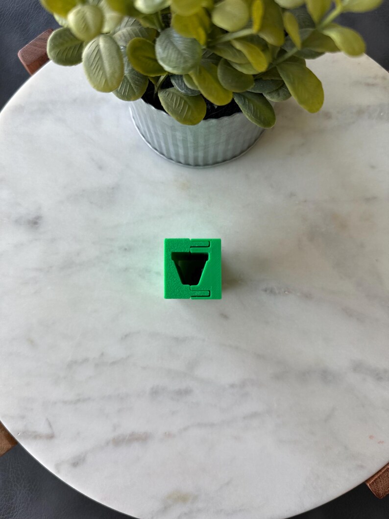 May include: A bright green, geometric cube with a V-shaped cutout. The cube is made of plastic and has a simple, modern design. The cube is sitting on a white marble surface.