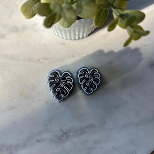 May include: Two black and white monstera leaf shaped earrings on a white marble surface.