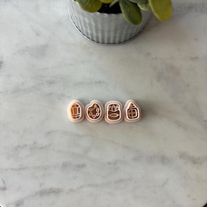 May include: Four beige cookie cutters in various shapes, including a house, a circle with dots, a face, and a gift box. The cutters are arranged in a row on a marble surface, with a small metal pot of greenery in the background.