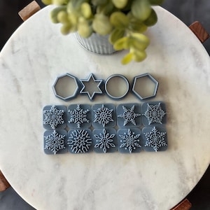 May include: A set of 12 snowflake-shaped silicone stamps, perfect for crafting and decorating. The stamps are arranged in three rows of four, with each stamp featuring a unique snowflake design. The stamps are a light blue color and have a white snowflake design.
