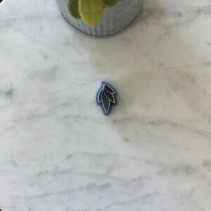 May include: A blue silicone stamp with a white outline of a leaf design. The stamp is on a white marble surface.