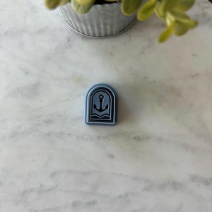 May include: A small, blue, rectangular mold with an anchor and wave design. The mold is set against a white and gray marble surface. A small, silver bucket with green foliage is in the background.