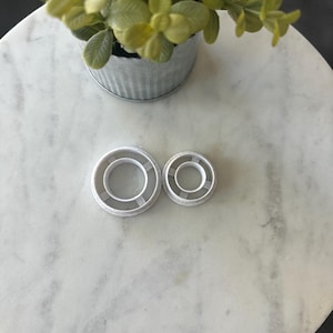 May include: Two white plastic cookie cutters in the shape of a circle with a smaller circle inside. The cutters are on a white marble surface.