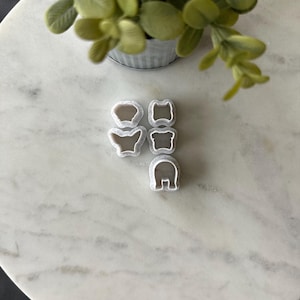 May include: Five white plastic cookie cutters in various shapes, including a bear, a butterfly, a heart, and two other shapes, on a white marble surface.