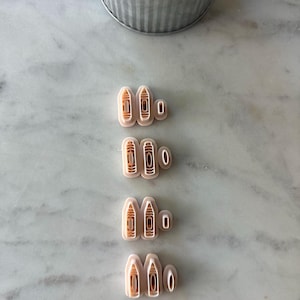 May include: A collection of four sets of beige-colored cookie cutters, each with a unique design. The cutters are arranged on a marble surface, with a small metal pot of greenery in the background. The cutters appear to be for making cookies.