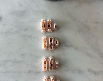 May include: A collection of four sets of beige-colored cookie cutters, each with a unique design. The cutters are arranged on a marble surface, with a small metal pot of greenery in the background. The cutters appear to be for making cookies.