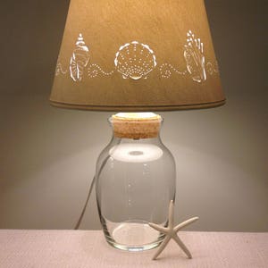 May include: A table lamp with a brown paper shade featuring a cut-out design of seashells and seaweed. The lamp base is a clear glass jar with a cork stopper.