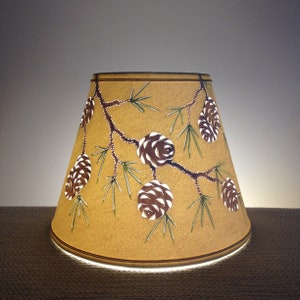 May include: A beige lampshade with a cut-out design of pine branches and pine cones. The pine cones are brown and white.