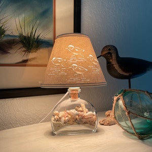 May include: A table lamp with a beige lampshade featuring a cut-out design of birds. The lamp base is a clear glass bottle filled with seashells and sand.