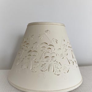 May include: Cream-colored lampshade with a floral cut-out design. The lampshade has a conical shape and a ribbon trim along the top and bottom edges. The cut-out pattern features stylized flowers and leaves.