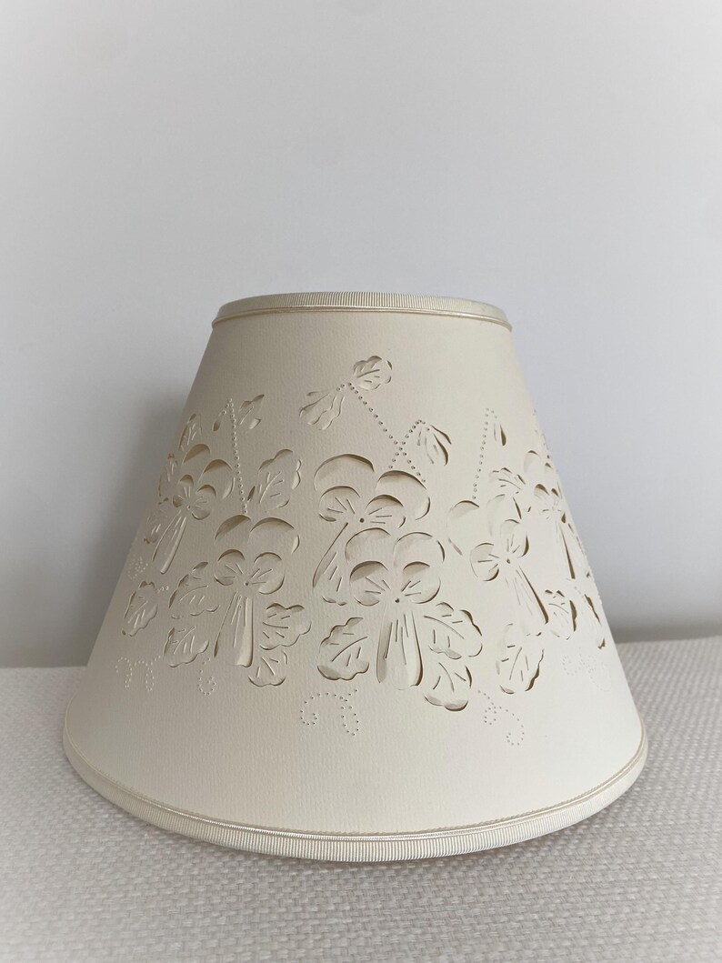 May include: Cream-colored lampshade with a floral cut-out design. The lampshade has a conical shape and a ribbon trim along the top and bottom edges. The cut-out pattern features stylized flowers and leaves.