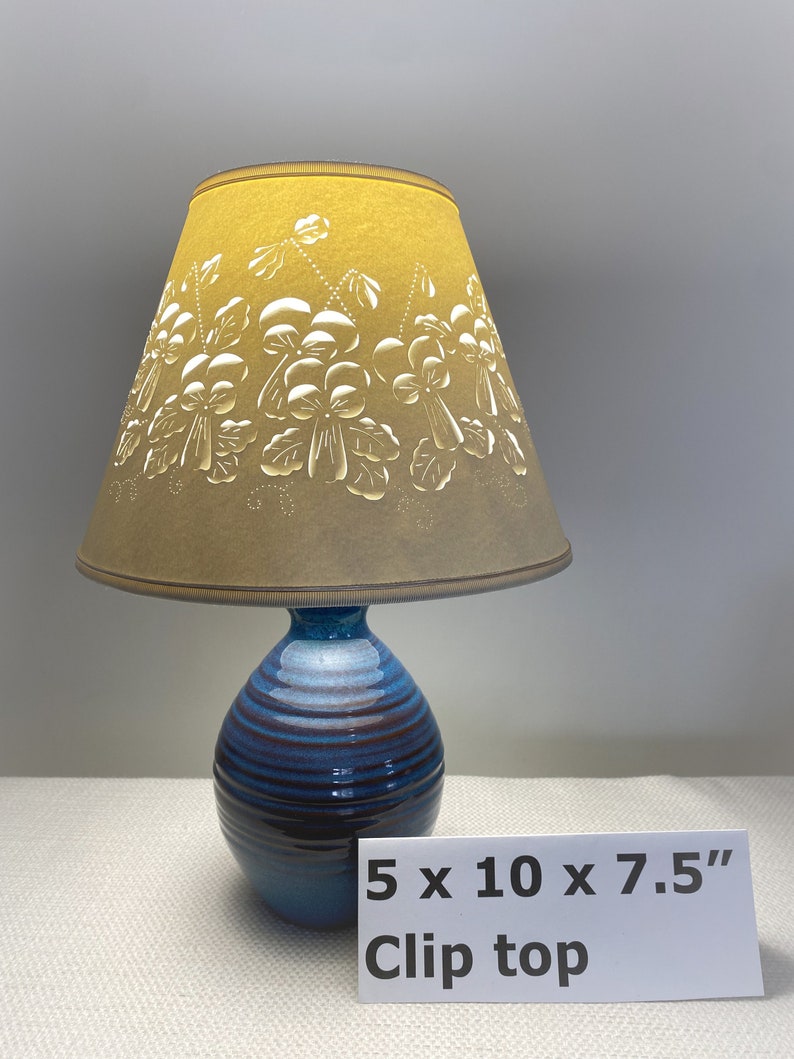 May include: A blue and brown ceramic table lamp with a clip-top lampshade. The lampshade is tan and features a floral design. The lamp base is 5 inches wide, 10 inches tall, and 7.5 inches deep. The text "Clip top" is visible.