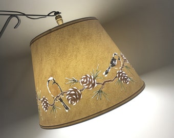 May include: A brown lampshade with a white bird and pine cone design. The lampshade is made of fabric and has a fringed edge.
