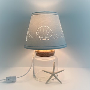 May include: A blue lampshade with a seashell design sits atop a clear glass jar with a cork lid. A white starfish sits in front of the jar.