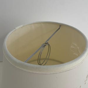 May include: Cream-colored lampshade with a decorative floral pattern. The lampshade has a cylindrical shape with a metal frame and a white trim around the top and bottom edges. The interior is a lighter cream color.