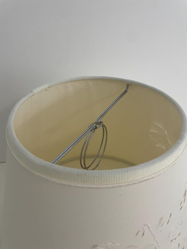 May include: Cream-colored lampshade with a decorative floral pattern. The lampshade has a cylindrical shape with a metal frame and a white trim around the top and bottom edges. The interior is a lighter cream color.