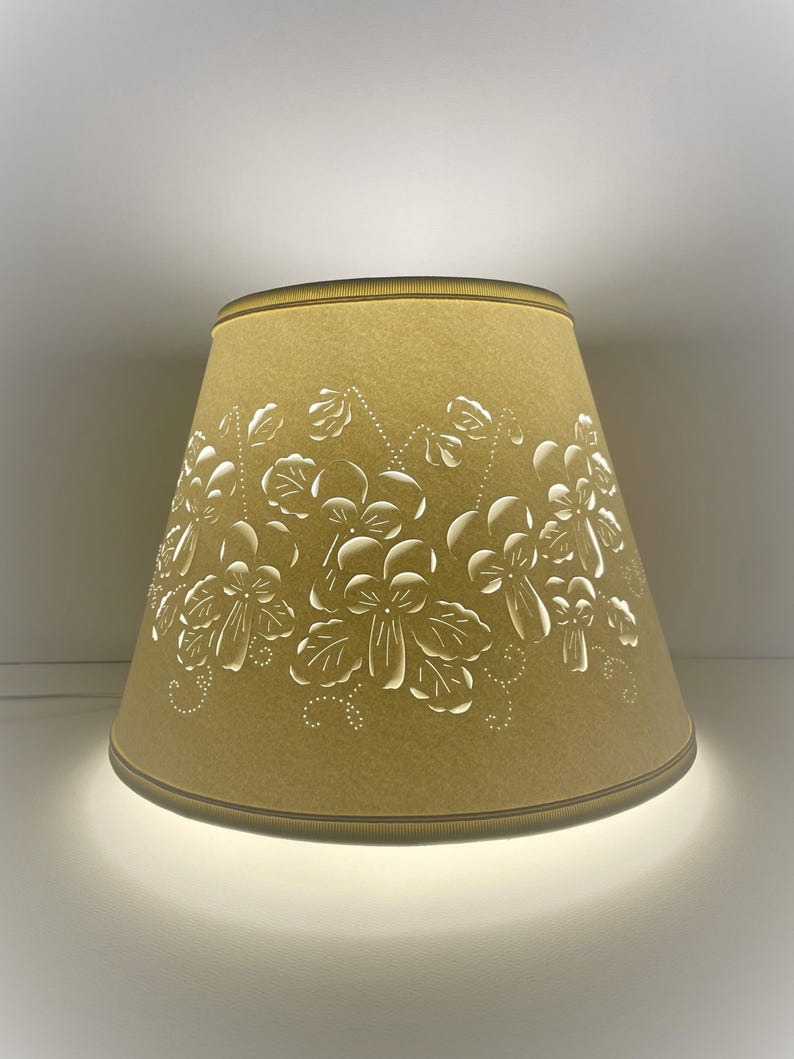 May include: A lampshade with a light tan color and a floral design cut out of the shade. The lampshade has a gold trim at the top and bottom. The light from the lamp shines through the cut-out floral design.