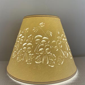 May include: A mustard-yellow lampshade with a floral design. The shade has a conical shape and is adorned with cut-out white flowers and leaves. The top and bottom edges are trimmed with a thin brown border. The light shines through the cut-out design.