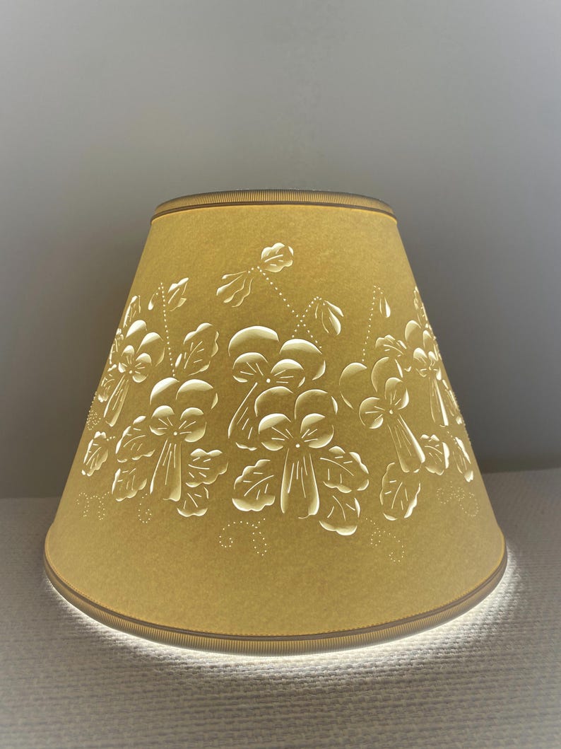 May include: A mustard-yellow lampshade with a floral design. The shade has a conical shape and is adorned with cut-out white flowers and leaves. The top and bottom edges are trimmed with a thin brown border. The light shines through the cut-out design.