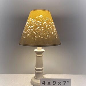 May include: A small table lamp with a mustard-colored lampshade featuring a floral design. The base is off-white and has a classic, turned design. The lamp is approximately 4 x 9 x 7 inches.