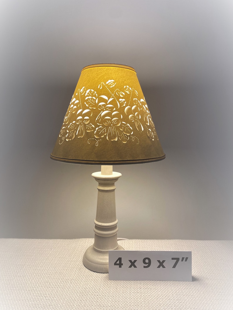 May include: A small table lamp with a mustard-colored lampshade featuring a floral design. The base is off-white and has a classic, turned design. The lamp is approximately 4 x 9 x 7 inches.