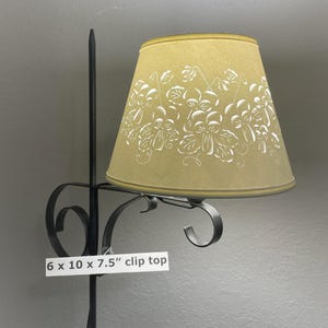 May include: A clip-top lampshade with a floral design. The shade is 6 inches wide, 10 inches long, and 7.5 inches tall. The shade is a light yellow color with white floral patterns. The lamp has a black metal frame.